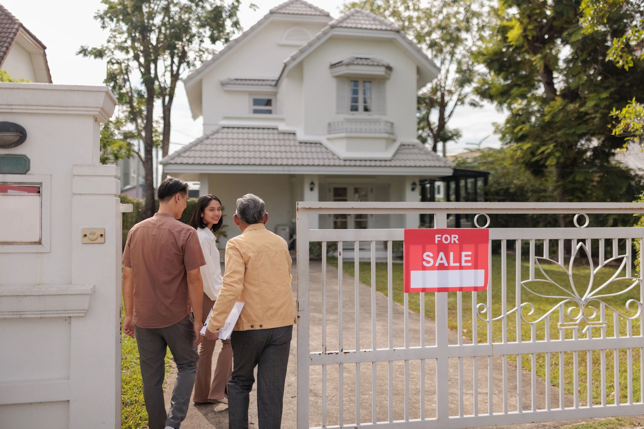 Couple with real estate agent walking through the gate of a white stucco home with a Spanish tile roof and for sale sign.