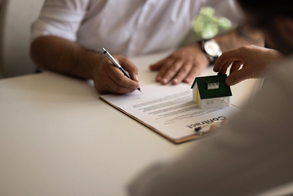 Man signing a real estate contract while another places a toy house on the paperwork.